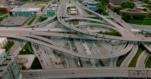 Aerial view of a complex highway interchange with multiple lanes and vehicles, surrounded by urban buildings and greenery.