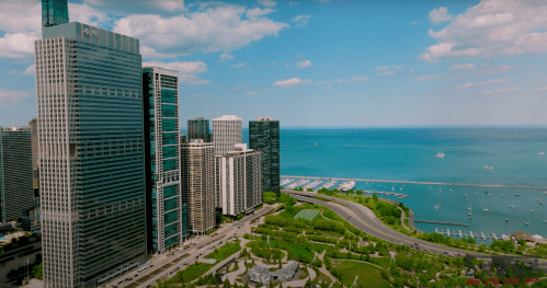 Aerial view of modern skyscrapers along a waterfront with boats and green spaces under a blue sky.