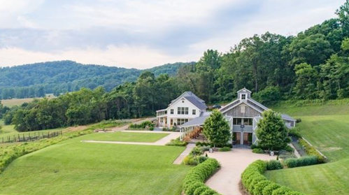 Aerial view of a scenic countryside property with two buildings, lush green lawns, and surrounding trees.