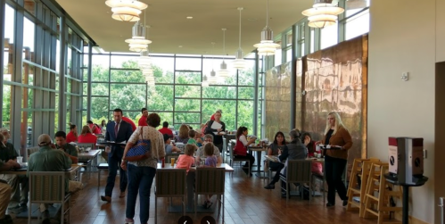 A busy café with large windows, people dining, and staff serving in a bright, modern space.
