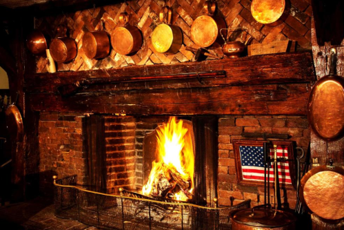 Cozy fireplace with a roaring fire, surrounded by rustic brick and copper cookware, featuring an American flag.