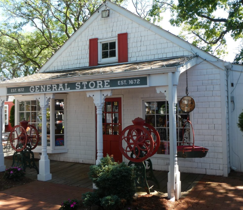 A charming white general store with red shutters, established in 1672, surrounded by greenery and colorful flowers.