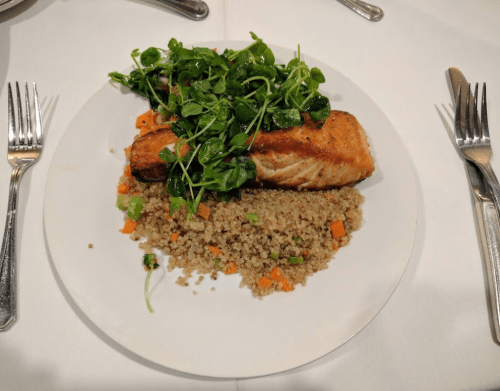 A plate with grilled salmon topped with greens, served over a bed of quinoa and diced vegetables.