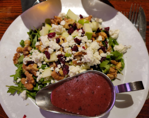 A fresh salad with greens, apples, walnuts, cranberries, and feta cheese, served with a side of berry dressing.