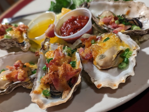 A plate of baked oysters topped with cheese, bacon, and green onions, served with lemon and dipping sauces.
