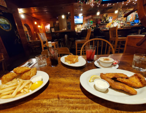 A wooden table set with plates of fried fish, fries, and drinks in a cozy restaurant with a bar in the background.