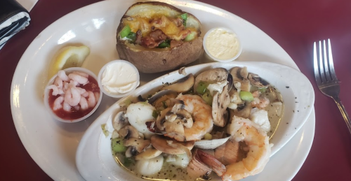 A plate of seafood with shrimp and mushrooms, served with a baked potato topped with cheese and bacon, and dipping sauces.
