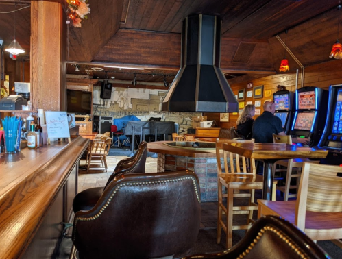 Interior of a cozy bar with wooden decor, a large hooded stove, and patrons at the bar and gaming machines.