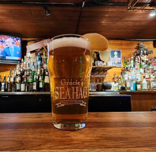 A pint of beer with a lemon slice sits on a wooden bar, with a well-stocked liquor shelf and a TV in the background.