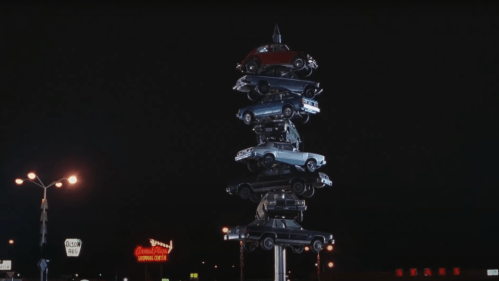 A tower of stacked cars illuminated at night, with a neon sign in the background.