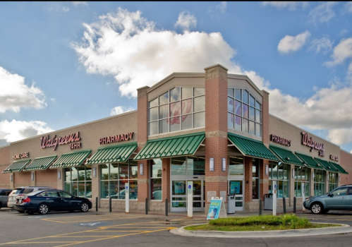 A Walgreens store with a green awning, featuring a pharmacy and drive-thru, set against a cloudy sky.