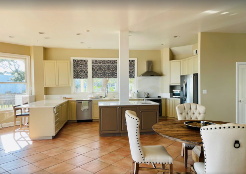 Bright, modern kitchen with beige cabinets, a large island, and a dining table, featuring tile flooring and large windows.