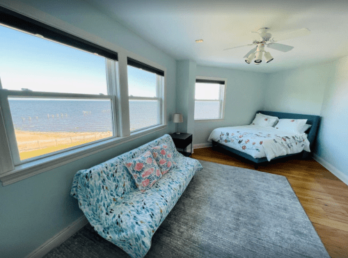 Bright bedroom with a view of the water, featuring a bed, a sofa, and large windows letting in natural light.