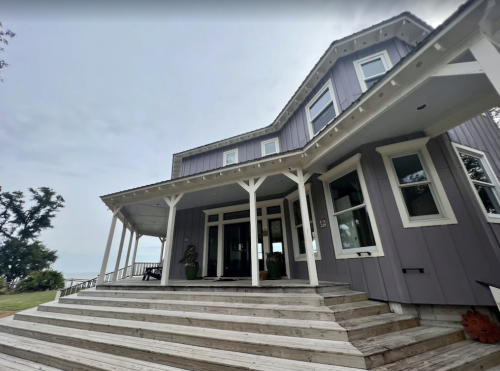 A large, modern house with a wraparound porch and multiple windows, set against a cloudy sky.