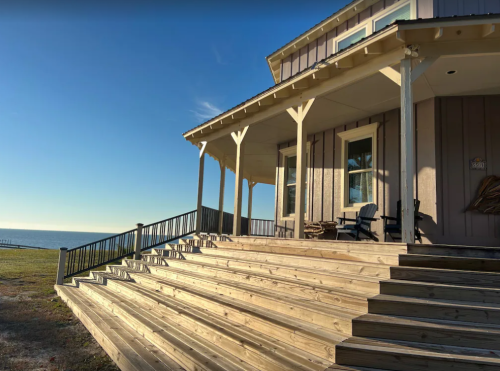 A wooden deck leads up to a house by the water, with clear blue skies and a calm sea in the background.