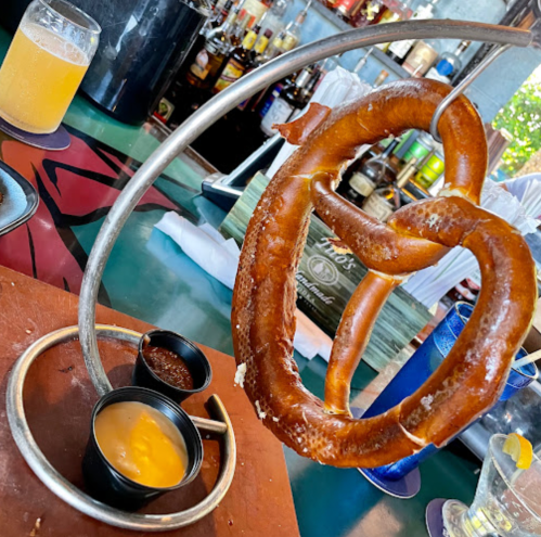 A large pretzel hanging from a metal stand, served with cheese and mustard dips on a wooden platter.