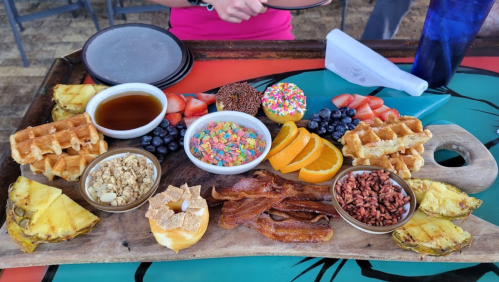 A colorful brunch platter featuring waffles, fruits, bacon, granola, and sweet treats on a wooden board.