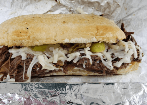 A sandwich filled with shredded beef, cheese, and pickles, served in a toasted hoagie roll, wrapped in foil.