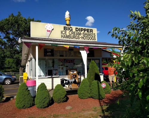 A small ice cream shop named "Big Dipper" with a sign, colorful flags, and greenery in front on a sunny day.