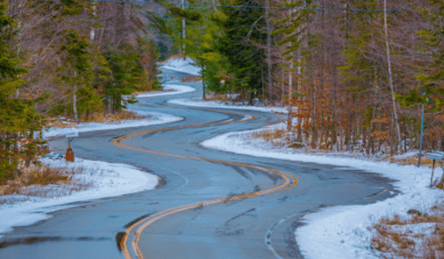 Winding road through a snowy landscape, surrounded by trees in autumn colors.