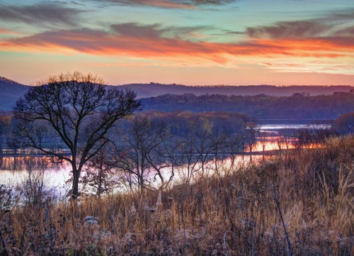 A serene landscape at sunset, featuring a river, hills, and silhouetted trees against a colorful sky.