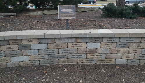 A stone wall with engraved names and a plaque, surrounded by trees and mulch in a park setting.