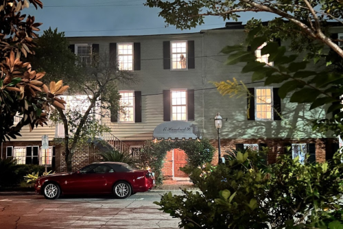 A charming two-story house at night, featuring lit windows, a red car parked outside, and lush greenery.