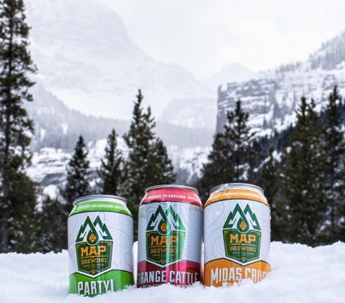 Three colorful beer cans from MAP Brewing sit in the snow, with a mountainous landscape in the background.