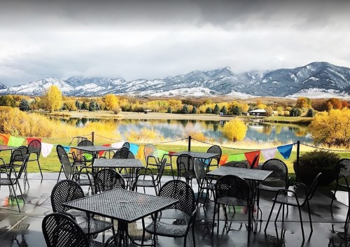 A scenic view of a lake surrounded by mountains, with empty outdoor tables and colorful flags in the foreground.