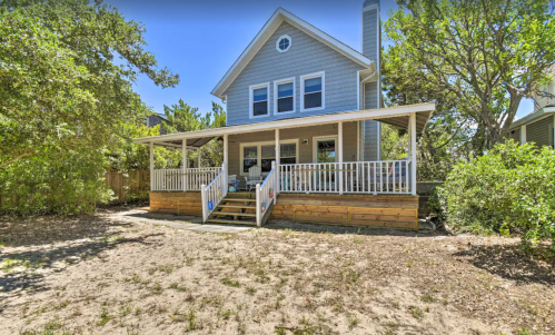 A two-story gray house with a front porch, surrounded by greenery and sandy ground.