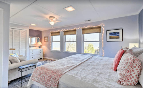 Bright bedroom with a large bed, cozy seating area, and windows adorned with soft lighting and scenic decor.
