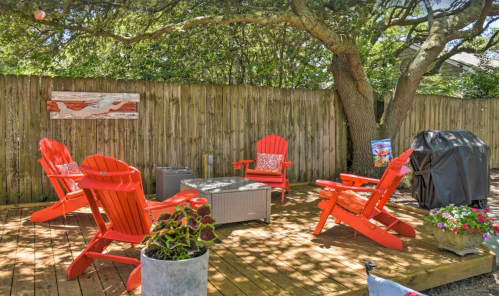 A cozy outdoor seating area with red Adirondack chairs, a table, and a grill, surrounded by greenery and a wooden fence.