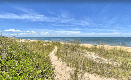 A sandy beach with gentle waves, framed by tall grass and a clear blue sky with scattered clouds.