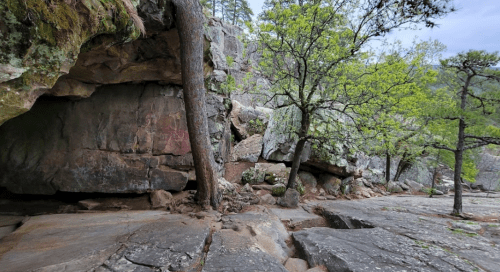 Rocky landscape with a cave entrance, surrounded by trees and rugged terrain under a cloudy sky.