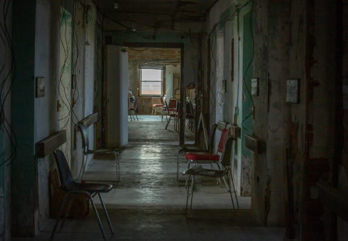 A dimly lit hallway with empty chairs and peeling walls, creating an eerie, abandoned atmosphere.