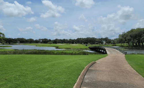 A scenic view of a golf course with a winding path, a bridge, and a serene lake under a partly cloudy sky.