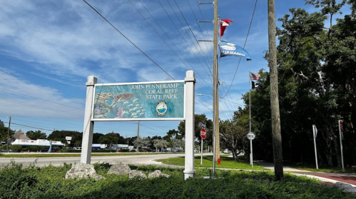 Sign for John Pennekamp Coral Reef State Park, with flags and trees in the background along a roadside.