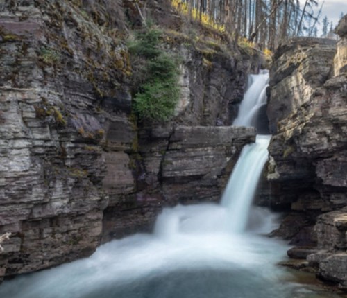 A serene waterfall cascades through rocky cliffs, surrounded by trees in a tranquil natural setting.