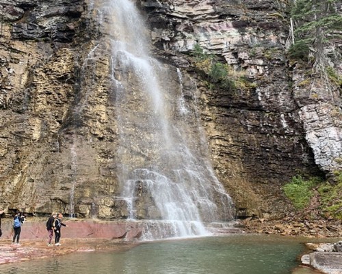 A waterfall cascading down rocky cliffs into a serene pool, with two hikers exploring the area nearby.