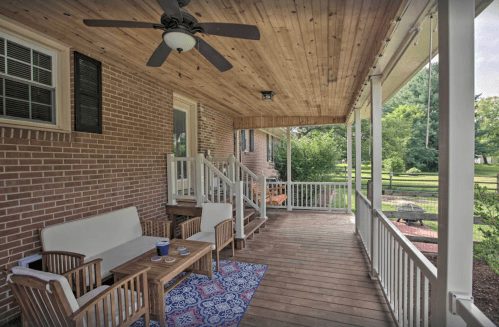 A cozy porch with a wooden ceiling, seating area, and a blue rug, surrounded by greenery and a peaceful view.