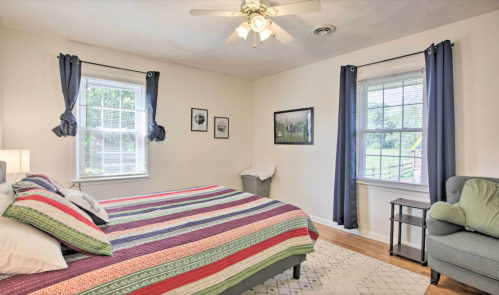Cozy bedroom with a striped bedspread, two windows, a chair, and framed pictures on the walls.