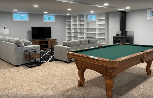 A spacious basement with a pool table, cozy seating area, and a TV, featuring light gray walls and carpet.