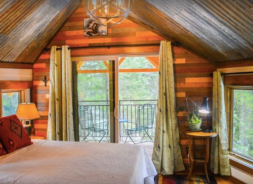 Cozy bedroom with wooden walls, a balcony view, and rustic decor, featuring a bed, lamp, and small table with a plant.