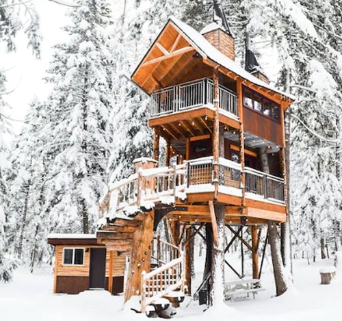 A tall wooden treehouse surrounded by snow-covered trees, featuring a staircase and a small cabin nearby.
