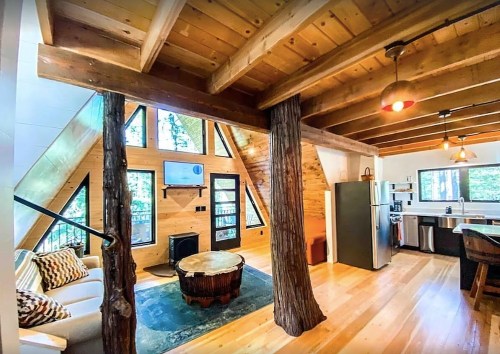 Cozy A-frame cabin interior with wooden beams, a fireplace, and modern kitchen, surrounded by large windows.
