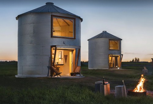 Two converted grain silos with open doors, set in a grassy field at dusk, with a campfire in front.