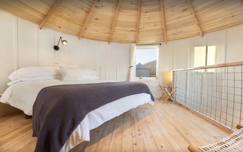 Cozy circular bedroom with wooden ceiling, large bed, and natural light from a window. Minimalist decor and warm tones.
