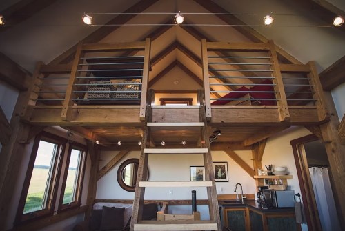 Cozy interior of a tiny house featuring a loft, wooden beams, and large windows with a staircase leading up.