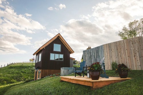 A cozy cabin on a grassy hill, featuring a wooden deck with chairs and potted plants, under a partly cloudy sky.