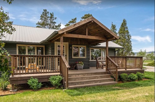 A cozy green house with a wooden porch, surrounded by greenery and trees under a clear blue sky.
